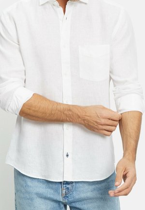 Uomo che si rimbocca le maniche di una camicia bianca con bottoni, indossando jeans azzurri davanti a uno sfondo semplice.