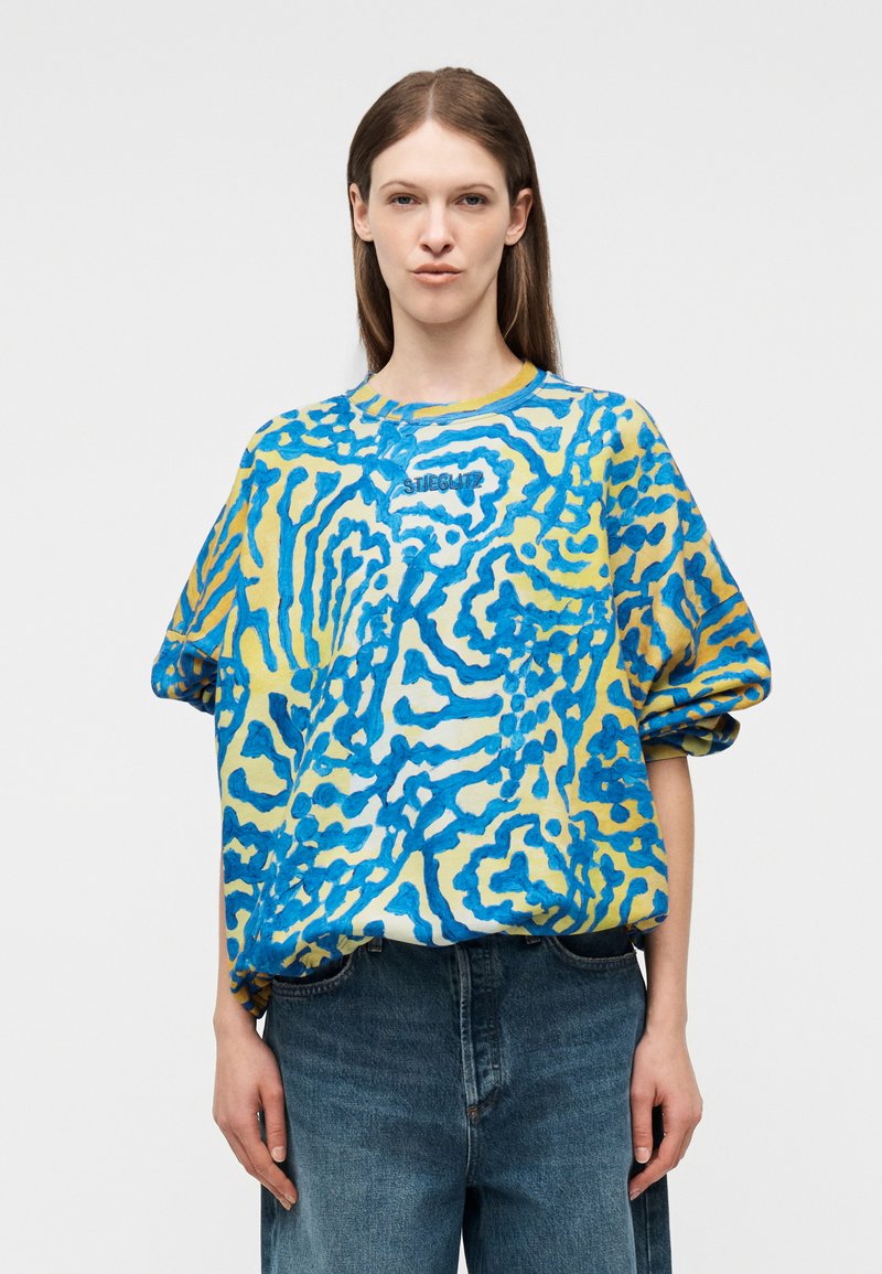 Νεαρό άτομο με ίσια καστανά μαλλιά φοράει φαρδιά μπλε και κίτρινα ρούχα με μακριά μανίκια και μπλε τζιν, στέκεται μπροστά σε απλό φόντο.