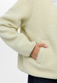 Veste en polaire crème avec une finition texturée, dotée d'une poche latérale, de poignets côtelés et d'une coupe décontractée.