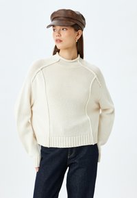 Maglione lavorato a maglia color crema con collo alto, orlo a coste e spalle cadenti, abbinato a jeans in denim scuri e un cappello in pelle marrone.