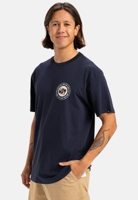 Camiseta de algodón azul marino de manga corta con un logo redondo en el pecho y un corte relajado. Presenta un cuello redondo y una etiqueta en el dobladillo lateral.