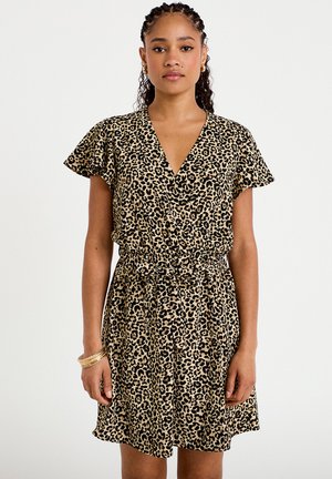 Femme aux cheveux tressés portant une robe à manches courtes imprimée léopard avec un col en V et une taille nouée, debout sur un fond blanc.