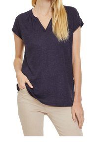 Námořnická modrá tričko s krátkým rukávem a V výstřihem, vyrobené z měkkého materiálu, s jemným nařasením na výstřihu a volným střihem. Nosené s světlými béžovými kalhotami.