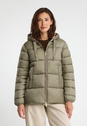 Donna che indossa una giacca trapuntata verde con cappuccio e coulisse e pantaloni beige chiaro, in piedi davanti a uno sfondo chiaro semplice.
