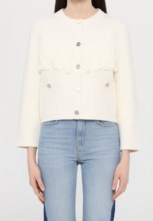 Femme portant un cardigan blanc boutonné avec des détails en franges sur la poitrine et un jean bleu clair taille haute.