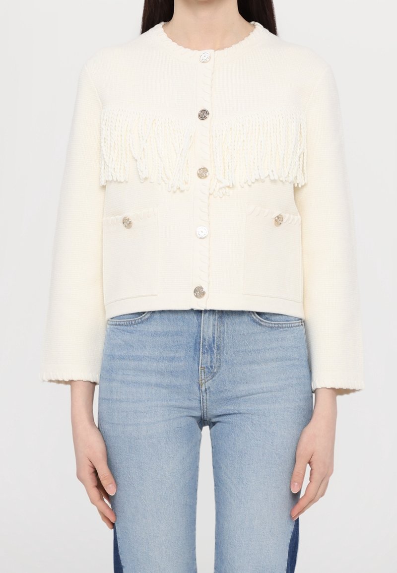 Femme portant un cardigan blanc boutonné avec des détails en franges sur la poitrine et un jean bleu clair taille haute.
