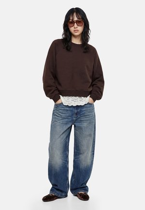Kvinde iført oversized brun sweatshirt, hvid top med blonder, løse blå jeans, brune flade sko og store brune solbriller, stående med hænderne i lommerne.