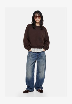 Kobieta w dużej brązowej bluzie, białym topie z koronką, luźnych niebieskich dżinsach, brązowych baletkach i dużych brązowych okularach przeciwsłonecznych, stojąca z rękami w kieszeniach.