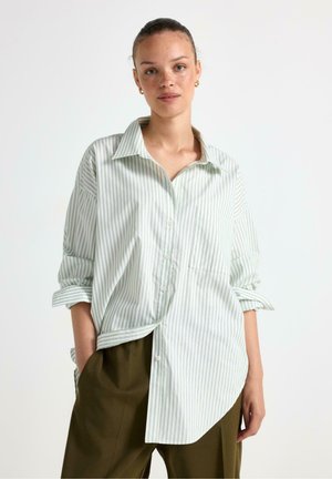 Camicia a righe verde chiaro e bianco con colletto, vestibilità ampia e una tasca sul petto, abbinata a pantaloni verdi scuri.