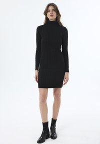 Frau in einem schwarzen kabelgestrickten Rollkragenkleid mit langen Ärmeln und schwarzen Stiefeletten, stehend vor einem weißen Hintergrund.