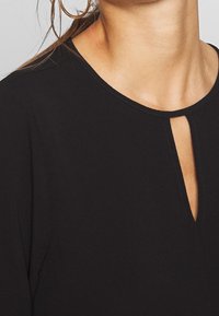 Top nero con scollatura tonda caratterizzata da un'apertura a chiave, maniche corte e una texture di tessuto liscia. Dettagli di cucitura sottili lungo i bordi.