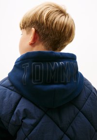 Námornícky prešívaný pufový kabát s kapucňou. Kapucňa obsahuje text "TOMMY" v tmavšej farbe. Hladký, textúrovaný materiál a diamantový vzor.