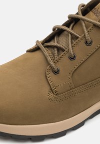 Brun mockaskinna ankelsko med texturerad mesh-tunga, runda snören och gummisula. Har metalöglor och en kontrasterande ljus sula.