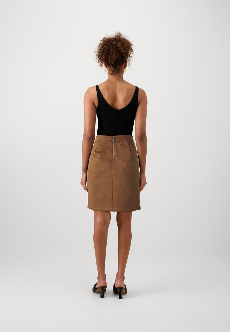 Femme aux cheveux bouclés portant un haut noir sans manches, une jupe en daim marron avec une fermeture éclair à l'arrière, et des sandales à talons noires, se tenant devant un fond blanc.