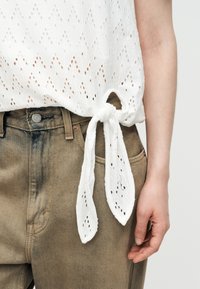 Λευκό τοπ από ύφασμα με σχέδιο eyelet και λεπτομέρεια δεσίματος στο πλάι, φορεμένο με ανοιχτό καφέ ψηλόμεσα τζιν και εμφανές χέρι που ακουμπά στο πλάι.