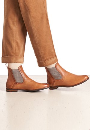 Stivali ankle in pelle marrone con pannelli in rete laterali, punta affusolata e cuciture decorative. Indossati con pantaloni in corduroy color sabbia.