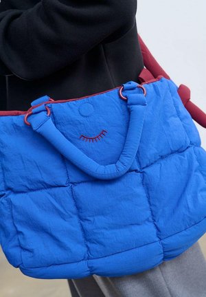 Niebieska pikowana torba na ramię z czerwoną lamówką, wyposażona w dwa uchwyty i przeszyty detal w kształcie uśmiechniętej buźki. Wykonana z miękkiej, teksturowanej tkaniny.