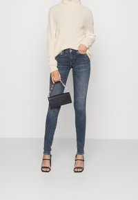 Donna che indossa un dolcevita crema, jeans skinny scuri, tacchi neri a strappo e tiene in mano una piccola borsa nera con una catena dorata.