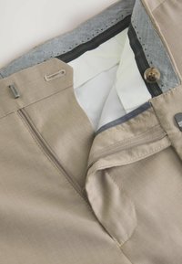 Tissu de pantalon beige avec une texture lisse, présentant une fermeture éclair, un bouton de fermeture et une taille à motifs. L'intérieur comprend une doublure blanche.