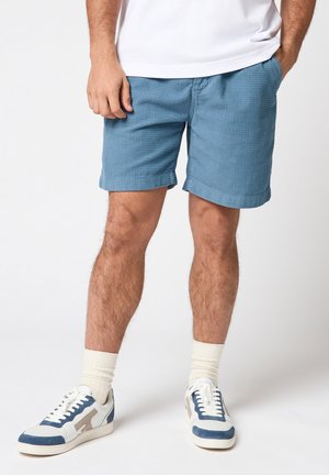 Homme portant un short bleu texturé, des chaussettes blanches, des baskets bleu et blanc, et une chemise blanche, debout avec une main dans la poche.
