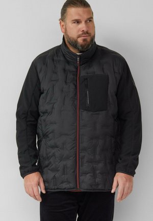 Mężczyzna z brodą i związanymi do tyłu włosami, ubrany w czarną pikowaną kurtkę z czerwonym zamkiem błyskawicznym i kieszenią na piersi, patrzący w bok.