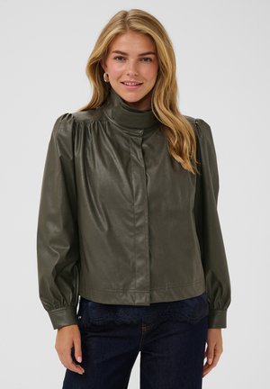 Frau mit langen blonden Haaren, die eine olivgrüne Lederjacke mit hohem Kragen und dunkelblaue Hosen trägt, leicht lächelnd vor einem schlichten Hintergrund.