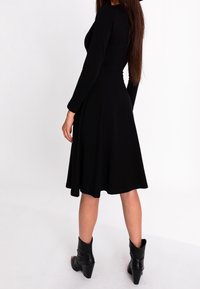 Femme portant une robe midi noire à manches longues avec une jupe évasée et des bottines noires à talons, se tenant devant un fond blanc.