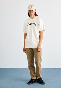 Camiseta color crema con logo "ellesse" en azul, pantalones beige claros y zapatos negros; gorra de color beige claro con logo; texturas suaves y corte casual.