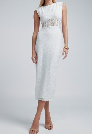 Femme portant une robe midi blanche sans manches en dentelle avec une partie centrale en dentelle transparente, des talons ouverts couleur nude et un bracelet en or au poignet droit.