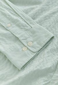 Poignet de chemise vert clair à manches longues avec deux boutons blancs et coutures visibles sur un tissu texturé.