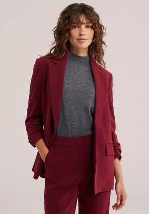 Donna con capelli mossi che indossa un completo bordeaux con maniche risvoltate e pantaloni abbinati su una maglia grigia a collo alto, mani rilassate.