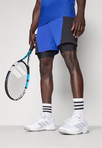 Camisa atlética azul, pantalones cortos negros y azules, zapatillas blancas con franjas grises y calcetines tobilleros a rayas; sosteniendo una raqueta de tenis negra.