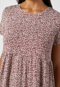 Vrouw in een jurk met korte mouwen, met een bijeen geraapte taille en een dicht patroon van kleine witte bloemen op een kastanjebruine achtergrond.