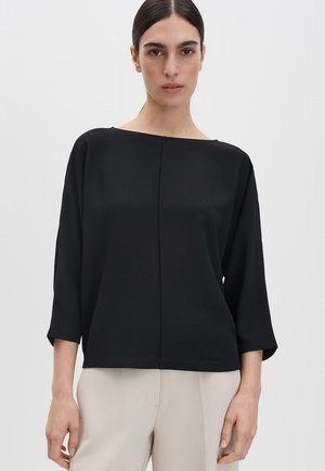 Woman with dark hair wearing a loose black long-sleeve top and light beige pants against a plain background.