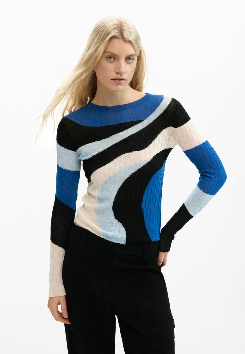 Junge Frau mit langen blonden Haaren, die einen eng anliegenden Pullover mit langen Ärmeln trägt, der abstrakte Farbblöcke in Blau, Schwarz, Weiß und Beige aufweist.