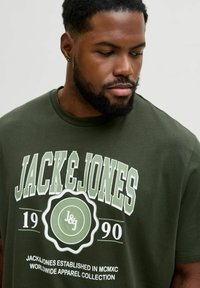 Zielona bawełniana koszulka z białym i jasnopodmiejskim nadrukowanym tekstem. Zawiera logo "JACK & JONES" oraz datę założenia, okrągły kształt graficzny.