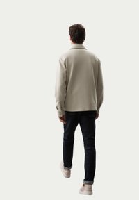 Camisa de manga larga beige claro, de corte casual, con cuello. Combinada con jeans azul oscuro y zapatillas de color claro. Hombre visto desde atrás.