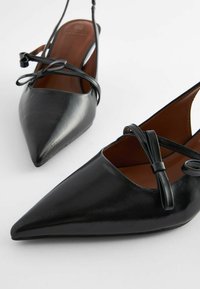Black leather slingback shoes with a pointed toe, featuring crossed straps with bows, smooth texture, and a brown insole.