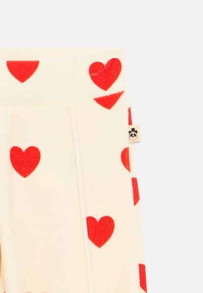 Miękka kremowa tkanina z czerwonymi wzorami serc i małą beżową metką z czarno-białą twarzą pandy na bocznej szwie.