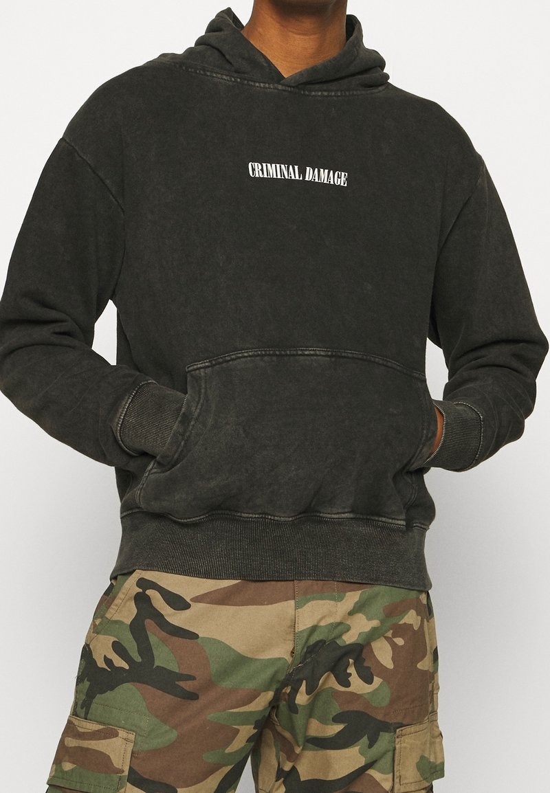 Personne portant un sweat à capuche noir délavé avec le texte "CRIMINAL DAMAGE" et un pantalon cargo camouflage, les mains dans la poche avant du sweat à capuche.