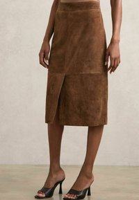 Gonna midi in suede marrone con uno spacco frontale, caratterizzata da linee pulite e una silhouette aderente. Indossata con tacchi aperti neri.