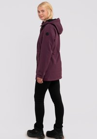 Lilla hættejakke med snørr i taljen, sidelommer og en sort detalje på armen, parret med sorte leggings og vandrestøvler.