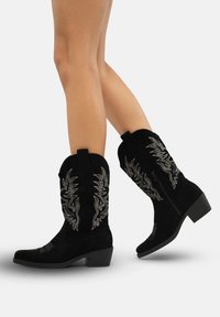 Las botas vaqueras de ante negro presentan bordados plateados, punta afilada, tacón bajo y lengüetas para facilitar su puesta.