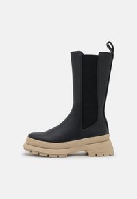 Czarny skórzany botek za kostkę z żebrowanymi, elastycznymi panelami po bokach, teksturowaną beżową gumową podeszwą oraz pętelką z tyłu ułatwiającą zakładanie.