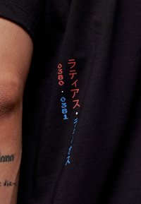 Zwarte katoenen T-shirt met multi-kleurige tekst in rood en blauw, met Japanse karakters en cijfers langs de mouw. Glad textuur.
