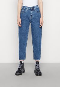 Žena v modrých džínech s vysokým pasem a černých šněrovacích botách, stojící na jednoduchém světlém pozadí.