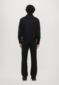 Černá mikina s kontrastním tyrkysovým prošíváním, vysokým límcem a přiléhavými manžetami, v kombinaci s černými kalhotami, hladká textura, uvolněná silueta.