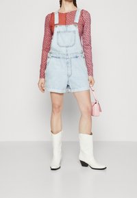 Denim-Latzshorts in heller Waschung mit verstellbaren Trägern, kombiniert mit einem roten, gemusterten, langärmeligen Oberteil und weißen Cowboy-Stiefeln. Rosa Handtasche inklusive.