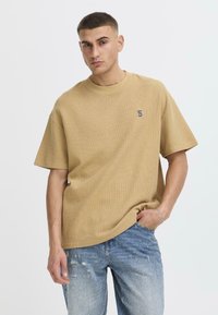 Camiseta de mangas cortas color marrón claro en un tejido texturizado, que presenta un pequeño logo negro en el pecho. Combinada con jeans azules desgastados.