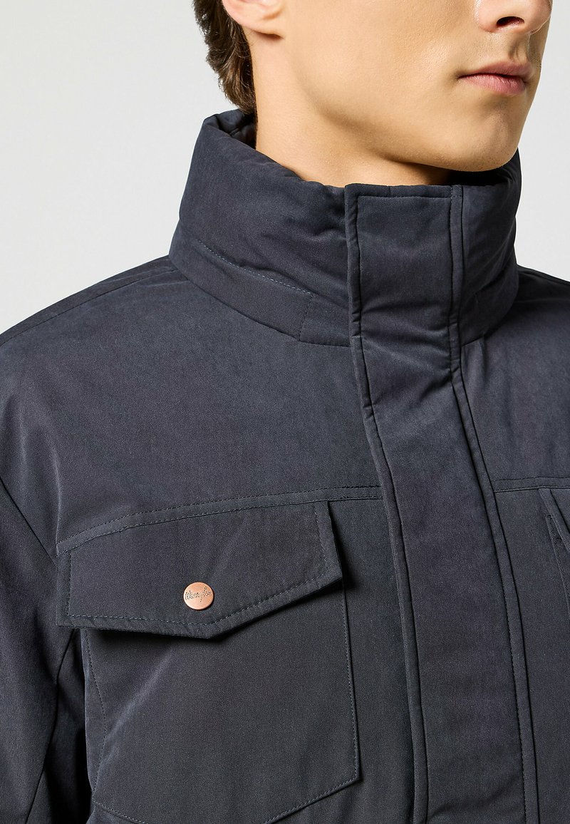 Giacca blu scuro con colletto alto, chiusura con zip e due ampie tasche sul petto con dettaglio a bottone color rame. Texture liscia.